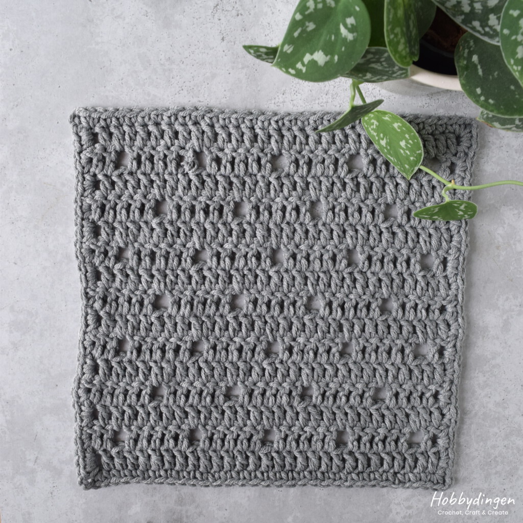 Grey crocheted square laying on a concrete looking background with a plant in the upper right corner.